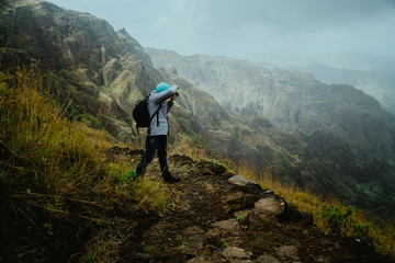 Naklejka na ściany i meble Hiker with camera in the steep mountainous terrain shooting a foto of lush canyon valley on the path from Xo-Xo Valley. Santo Antao Island, Cape Verde