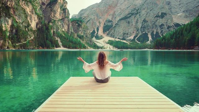 Young happy woman tourist sitting on wooden pier of Alpine Lake Braies, white sexy silk boho blouse turned away raises hands prays divine enjoy beautiful Italian mountains picturesque wild life nature
