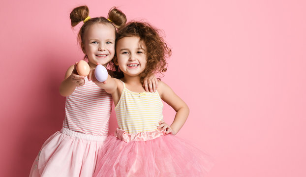 Two small smiling girls in pink stylish clothes standing and holding colored eggs in hands over pink background