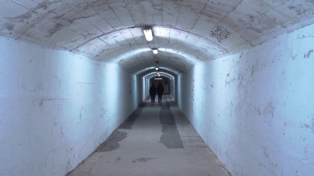 Two girls in jackets go down a long empty corridor or an underground pedestrian crossing. An old abandoned bunker or bomb shelter with dim lighting, peeling plaster, and graffiti on walls and ceiling.