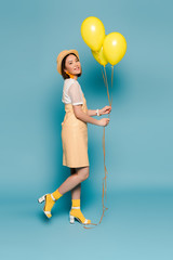 side view of happy asian girl in striped dress and straw hat with yellow balloons on blue background