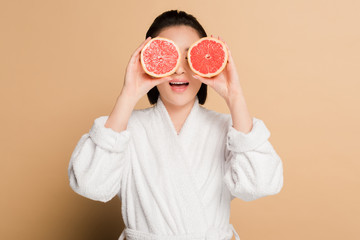 shocked beautiful asian woman in bathrobe with grapefruit halves on eyes on beige background