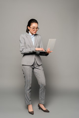 young businesswoman in eyeglasses using laptop on grey background