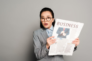 shocked young businesswoman in eyeglasses holding business newspaper on grey background