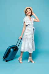smiling brunette asian girl in striped dress and straw hat with travel bag on blue background