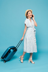 smiling brunette asian girl in striped dress and straw hat with travel bag talking on smartphone on blue background