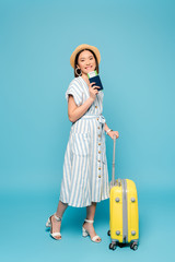 smiling brunette asian girl in striped dress and straw hat with travel bag and passport with air ticket on blue background