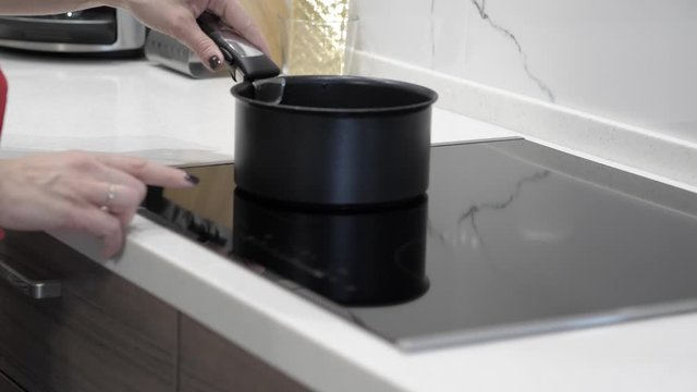 Female hands put a black stewpan on an induction stove and placed a silicone spatula inside, in the background a white kitchen