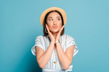 Fototapeta premium brunette asian girl in striped dress and straw hat touching face and pouting lips on blue background