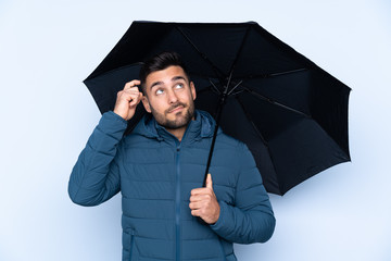 Man holding an umbrella over isolated background having doubts and with confuse face expression