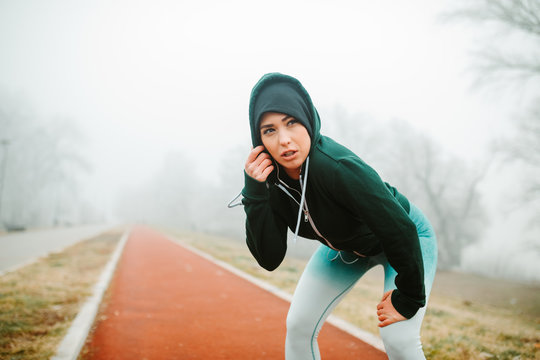 Young Woman Athlete Is Listening To Music On Her Headphones While Taking Break From Running. Fit Girl Is Trying To Take A Breath After Running On Cold And Foggy Day.