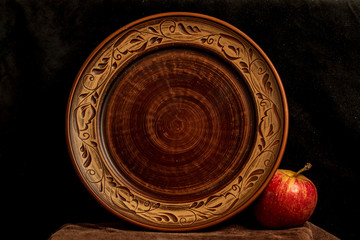 Empty clay plate with a pattern on a leather stand. Nearby lies a red apple. Black background. Direct look.