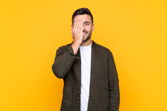 Man over isolated yellow background covering a eye by hand