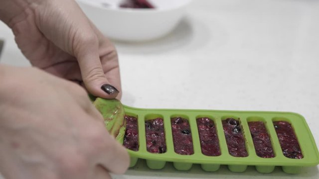 Female hands pull cubes from frozen berries from a green silicone mold and place them in a white cup.