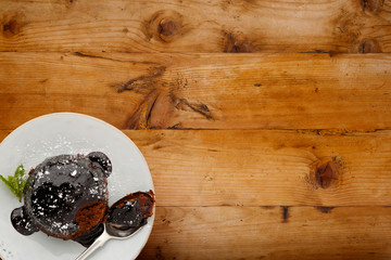 An individual chocolate sponge cake, with a sticky chocolate sauce, on a white plate with a spoon, shot on a wooden background with copy space.