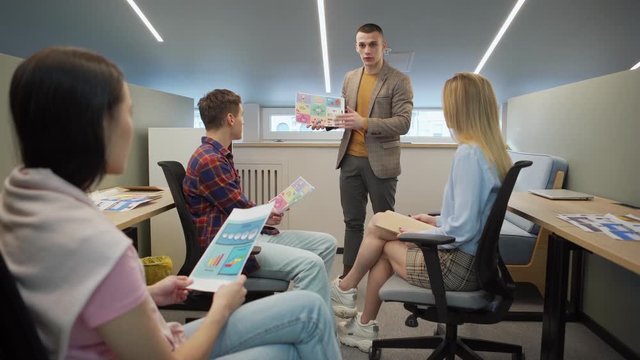 Young team leader presenting project idea to colleagues in small office. Manager showing paper plan and explaining details, teammates asking questions