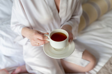 The girl is holding a white tea Cup and saucer. In white coats. Breakfast in bed. The morning starts with Breakfast. Hot tea in the hands of a young girl in a white coat sitting on a white bed