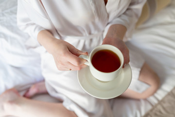 The girl is holding a white tea Cup and saucer. In white coats. Breakfast in bed. The morning starts with Breakfast. Hot tea in the hands of a young girl in a white coat sitting on a white bed