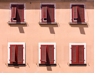 Fassade eines Hauses im mediterranen Stil, Putz sandfarben gestrichen mit geschlossenen Holzladen in dunkelrot, teilweise ausgestellt an 6 Fenstern während harter Mittagssonne mit kräftigen Schatten