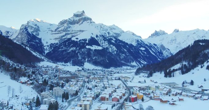Aerial shot of mountain village surrounded by Swiss Alps. Mountains partially covered in snow. Pine trees on mountain slopes. Switzerland. Drone.