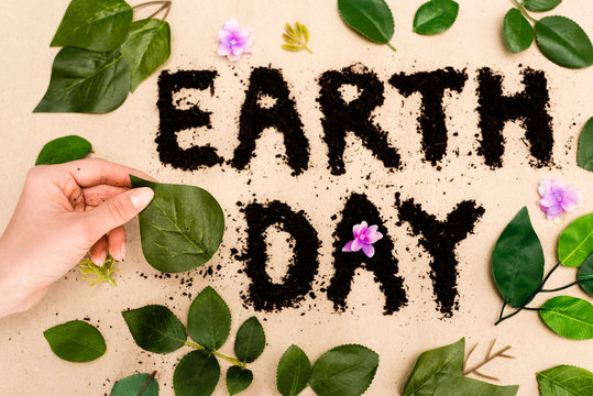 Cropped view of woman hand with leaves, buds and earth day lettering on beige