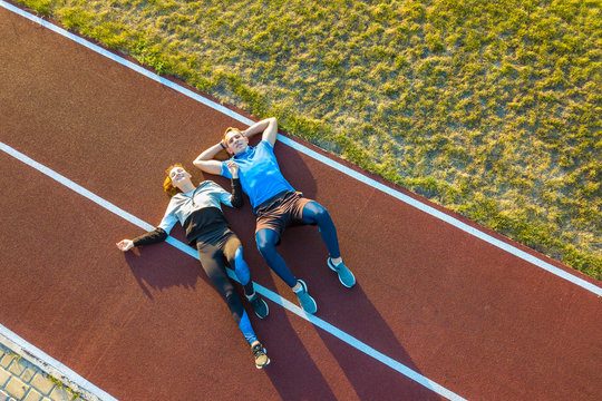 Top Down Aerial View Of Two Young People Sportsman And Sportswoman Laying On Red Rubber Running Track Of A Stadium Field Resting After Jogging Marathon In Summer.