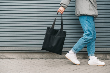 Closeup,cropped photo,man in stylish clothes goes on a background of a gray wall with an eco bag in a black bag in hand. Guy's legs walking against a wall background with a shopping bag. Copy space