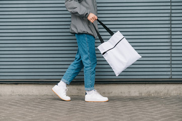 Cropped photo, stylish young man in street clothes walking with reusable shopping bag on gray background, legs and eco bag close up. copy space