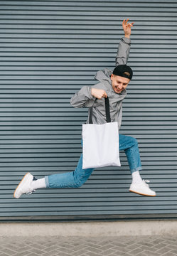 Happy Young Man In Stylish Casual Clothes Jumping On Gray Wall Background With Reusable Eco Bag In Hand On Gray Wall Background. Smiling Guy In Jump With Shopping Bag. Vertical