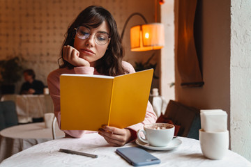 Pretty brunette young woman indoors in cafe reading book.