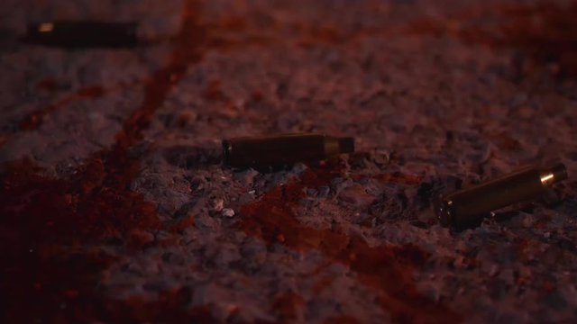 A slow sliding shot shows empty bullet casings strewn across asphalt, with red and blue police lights lighting up the scene.