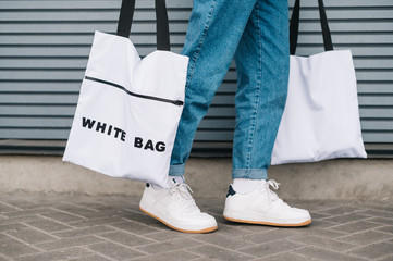 Eco friendly man carries 2 trendy reusable eco bags in his hands. Man legs in jeans and sneakers and 2 white stylish eco bags on a gray wall background, closeup photo, copy space. Background.