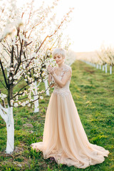 Obraz na płótnie Canvas Full-length portrait of young Caucasian woman with short blond hair in elegant light pink wedding dress is standing in a blooming spring fruit garden.