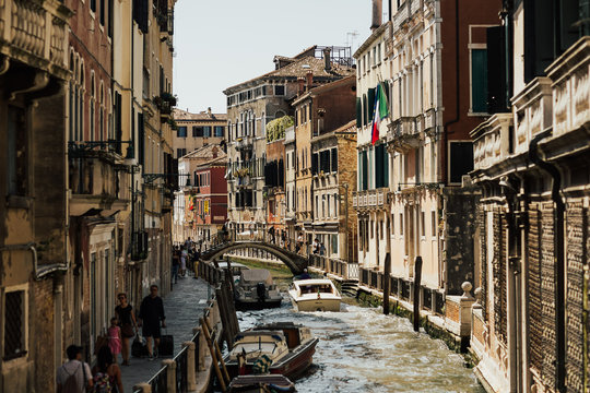 VENETIA, ITALY, JULY 17, 2019 Boats in Venetian harbor in front of houses. Old houses and bridge on the channel in Venice (Italy) on a sunny day in summer. Many tourists. Perfect for a romantic trip.