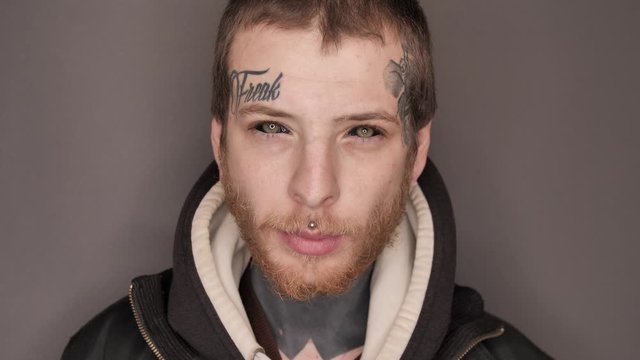 Close-up of a man with tattoo on his face and neck, eyeball or eye scleral tattooing, piercing, posing in studio, provocative aggressive look. Staring at the camera. Isolated on grey background.