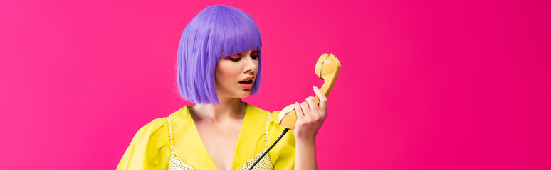 panoramic shot of beautiful emotional girl in purple wig holding retro telephone, isolated on pink