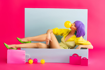 pop art girl in purple wig as doll blowing bubble gum while sitting in blue box with balls and shopping bags, on pink
