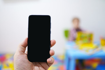 Parent holding smart phone on the background of child playing at a table in the nursery