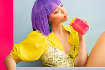 beautiful pop art girl in purple wig as doll drinking from jar while sitting in blue box, isolated on pink