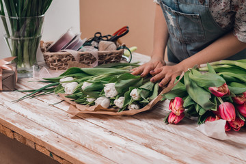 Hands of young woman florist working with fresh flowers making bouquet of pink tulips on table.