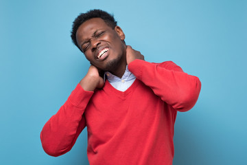African american young man in red sweater having terrible pain in neck suffering form spasm. Studio shot on blue wall