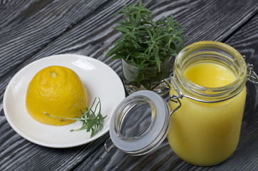 Lemon Kurd in a glass jar. Nearby is half a lemon on a saucer and a bunch of rosemary. Against the background of brushed boards.
