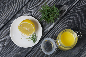 Lemon Kurd in a glass jar. Nearby is half a lemon on a saucer and a bunch of rosemary. Against the background of brushed boards.