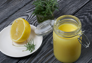 Lemon Kurd in a glass jar. Nearby is half a lemon on a saucer and a bunch of rosemary. Against the background of brushed boards.