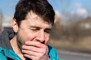 Fototapeta premium Coughing, sneezing or yawning squinting bearded man, covering his mouth with his hand on the street on a sunny day. Sick or tired man. Closeup view. Blurred background