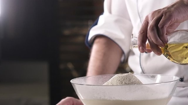 chef in the kitchen pours olive oil from a bottle in a thin stream into wheat flour. Preparing ingredients for baking delicious cookies or pasta. Preparation of dough in a pastry shop. Slow motion.