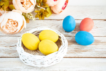 Fototapeta na wymiar Easter card. Pastel yellow eggs in a white wicker basket on a wooden white table. Flowers in the background. Copy space for text.