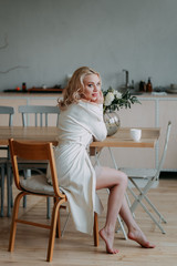 Bride's bouquet. Beautiful happy happy girl blonde bride in a white light sexy robe in the morning in the kitchen is happy, smiling and touching a bouquet of fresh flowers , preparing for the wedding