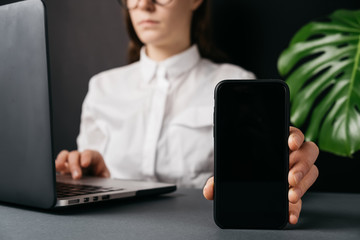 Cropped portrait of successful businesswoman in white shirt, showing blank copy screen cell phone for your information or advertisement, sitting at desk near pc. Selective focus