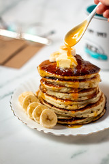 Hot american pancakes with with apricot jam dripping from the spoon. Served with bananas and melted butter on the white table. Mug, glasses and envelope background